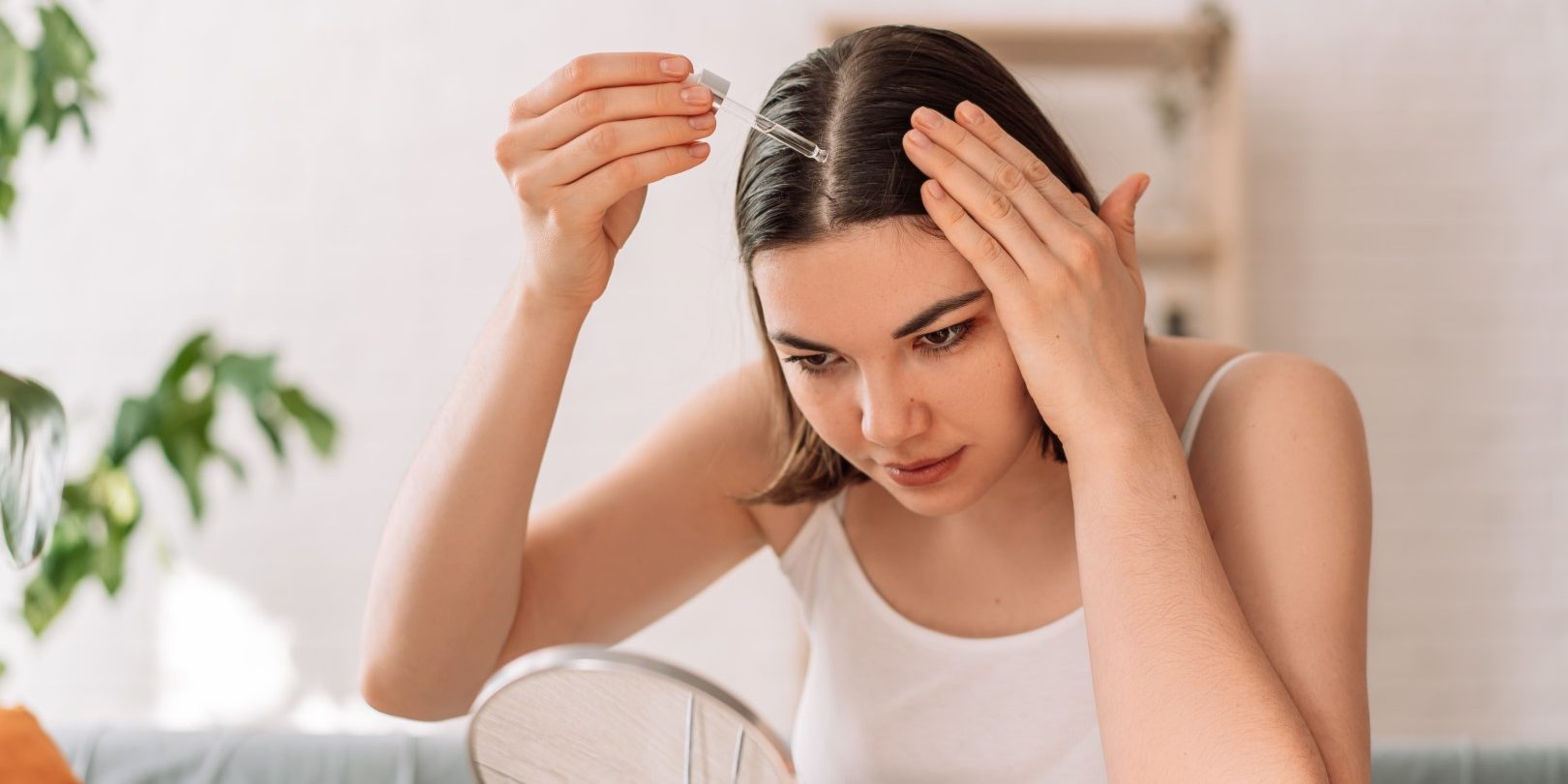Lady sitting on the couch looks in the mirror and drips hair oil with a pipette on her hair